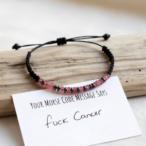 Pode incluir: Uma pulseira preta trançada com contas rosa e pequenas contas de metal preto. Um cartão branco com o texto "YOUR MORSE CODE MESSAGE SAYS" e "fuck Cancer" está abaixo da pulseira.