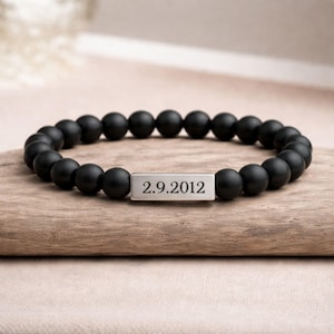 May include: A black beaded bracelet featuring a silver-coloured rectangular charm engraved with the date "2.9.2012". The bracelet is displayed on a piece of wood.