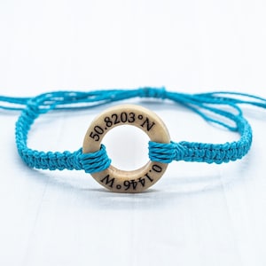 May include: A turquoise blue braided bracelet with a wooden circle charm that has the coordinates "50.8203&deg; N 0.1446&deg; W" engraved on it.