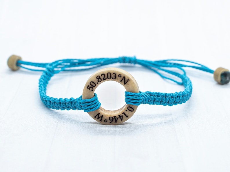 May include: A turquoise blue braided bracelet with a wooden circle charm that has the coordinates "50.8203&deg; N 0.1446&deg; W" engraved on it.