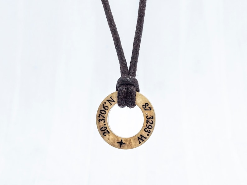 May include: A gold-colored necklace with a round pendant featuring the coordinates "20.3706&deg; N, 87.3293&deg; W" engraved on it. The necklace is made of a black cord.