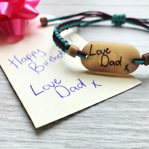 以下が含まれることがあります： ターコイズとマルーンのブレードブレスレット。木製タグには「Love Dad x」と刻まれています。ブレスレットは、「Happy Birthday Love Dad x」と青いインクで書かれた白いカードの上に置かれています。