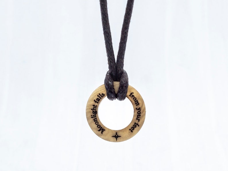 May include: A wooden ring necklace with a black cord. The ring has the words "Moonlight falls from your feet" engraved on it.