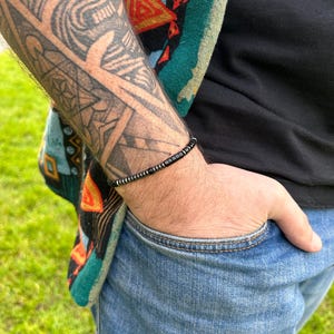 May include: A black beaded bracelet with silver accents. The bracelet is worn on the wrist of a person with a tattoo.