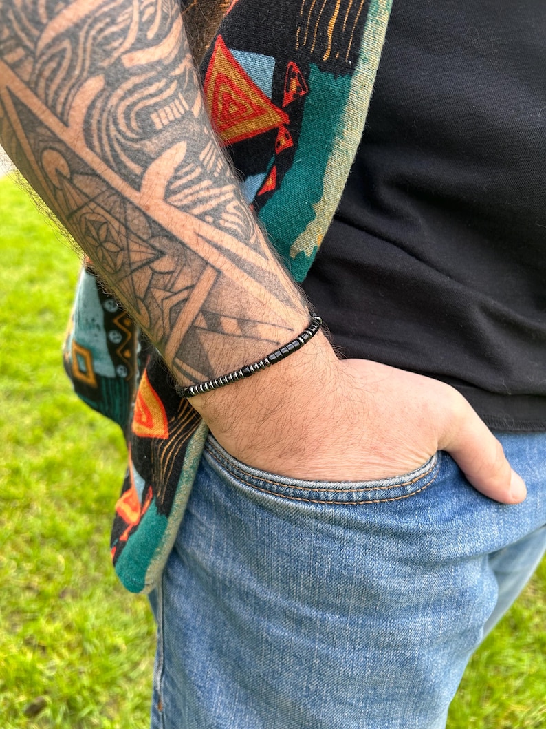 May include: A black beaded bracelet with silver accents. The bracelet is worn on the wrist of a person with a tattoo.
