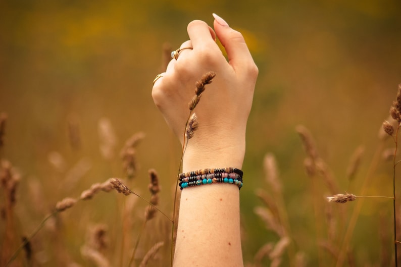 Three personalised beaded Morse code bracelets, made from black, natural and colourful beads. Pictured on ladies wrist with corn crop background.