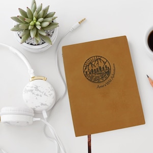 A brown, personalised journal stylishly arranged on a table. The faux leather cover is engraved with a unique travel notebook design, along with your name and the phrase “wild adventures”.