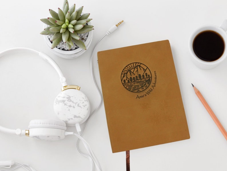 A brown, personalised journal stylishly arranged on a table. The faux leather cover is engraved with a unique travel notebook design, along with your name and the phrase “wild adventures”.