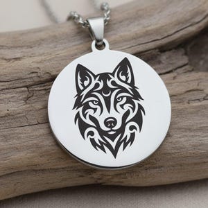 May include: A silver-colored pendant necklace featuring a detailed black wolf head design. The wolf's face is rendered in a tribal style, set against a circular backdrop. The necklace is displayed on a piece of driftwood.