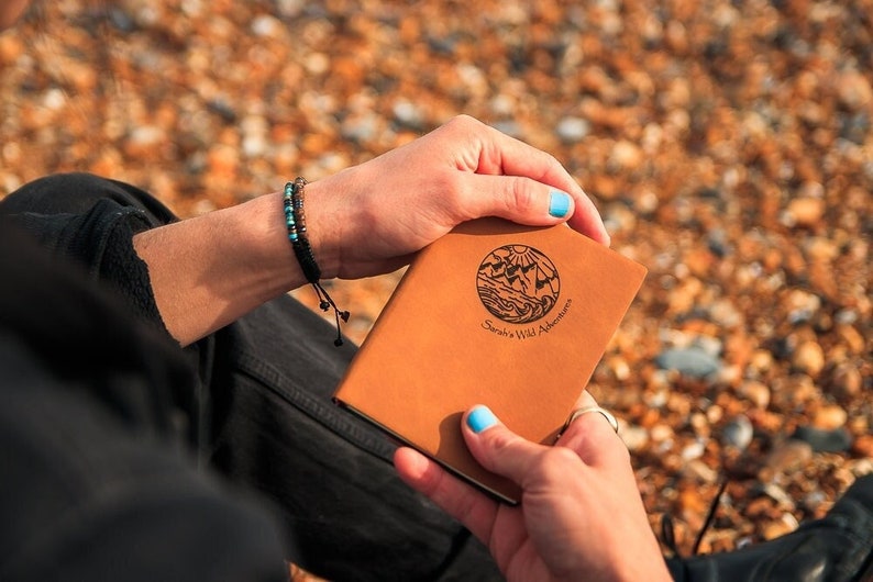 A brown, personalized journal being held in a hand. The cover is embossed with a unique travel design, along with your name and the phrase “wild adventures”.