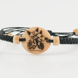 Puede incluir: Pulsera trenzada negra con un dije redondo de madera que presenta una ilustración en blanco y negro de dos zorros acurrucados.