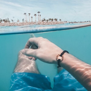 May include: A person's hand and arm are submerged in clear blue water, with a black bracelet with a gold circle charm. The water is shallow and the bottom is visible. The person is wearing blue swim trunks. In the background, a tropical island with palm trees is visible above the waterline.