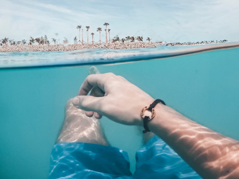 May include: A person's hand and arm are submerged in clear blue water, with a black bracelet with a gold circle charm. The water is shallow and the bottom is visible. The person is wearing blue swim trunks. In the background, a tropical island with palm trees is visible above the waterline.