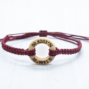 May include: A maroon braided bracelet with a wooden circle charm engraved with the coordinates "50.8203&deg; N 0.1446&deg; W".