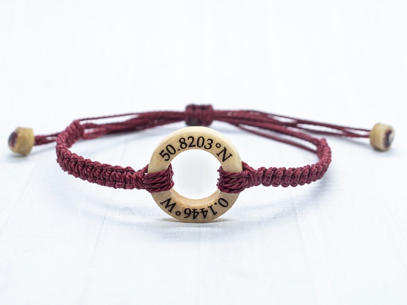 May include: A maroon braided bracelet with a wooden circle charm engraved with the coordinates "50.8203&deg; N 0.1446&deg; W".