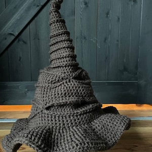 May include: A brown crocheted hat shaped like the Sorting Hat from the Harry Potter series. The hat has a pointed, curled top and a wide brim.