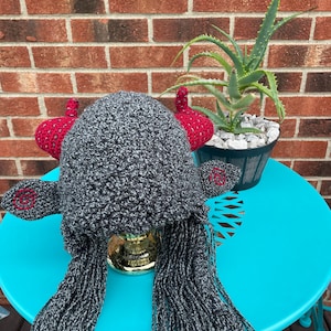 May include: A gray and black crocheted hat with red horns and long, flowing hair. The hat is sitting on a teal table.