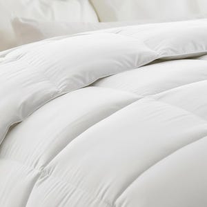 May include: Close-up of a white comforter with a quilted design. The comforter is made of a soft, fluffy material and is neatly arranged. The bedding is a solid white color, creating a clean and inviting look. The image suggests a focus on comfort and quality.