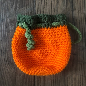 May include: A handmade orange crochet pouch with a green drawstring and top. The pouch is round and has a green handle. The pouch is sitting on a wooden surface.