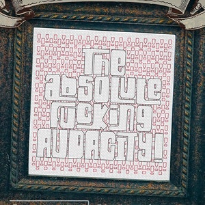 May include: A framed cross-stitch design with a white background and red squares. The design reads "The absolute fucking audacity!" in bold white letters. The frame is dark green with gold accents and a white banner at the top reads "Audacity" in a decorative font. The bottom of the frame reads "Urban Stitches" in white letters.