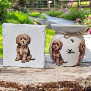 May include: A white square coaster and a white ceramic oil diffuser featuring a brown and black puppy illustration. The diffuser is labelled "Olive". The items are on a wooden surface.