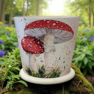 May include: A white ceramic plant pot with a saucer, decorated with a hand-painted design of two red and white spotted mushrooms. The pot has a speckled finish and sits on moss, with a blurred forest background.