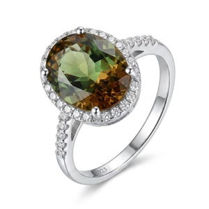 May include: A silver ring with a large, oval-cut gemstone. The gemstone has a green and brown color gradient, surrounded by a halo of small, clear stones. The ring band is also adorned with small stones.