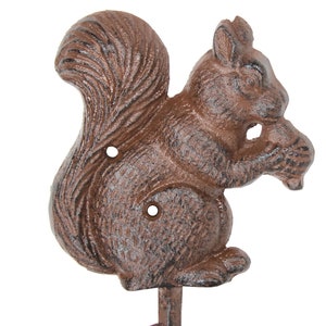 May include: A cast iron squirrel-shaped wall hook in a rustic brown finish. The squirrel is depicted holding a nut, with a curved tail and two mounting holes. The hook is a decorative and functional item.