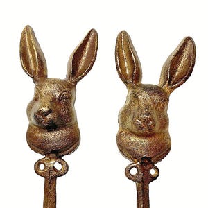 May include: Two bronze-colored metal wall hooks. Each hook features a rabbit head design with long ears and a textured finish. The hooks have a keyhole-shaped detail and a rounded hook at the bottom, perfect for hanging items.
