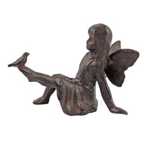 May include: A cast iron fairy figurine in a seated pose with outstretched leg and delicate wings. The fairy is finished in a dark brown color, with details in the wings and clothing. This decorative item is suitable for indoor or outdoor use.