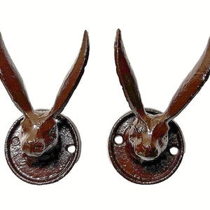 May include: Two identical wall-mounted hooks, each featuring a brown, textured cast metal hare head with long, upright ears. The hare head is attached to a circular, flat base plate that includes two pre-drilled holes for mounting purposes.