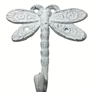 May include: A white, cast metal wall hook shaped like a dragonfly. The insect features textured wings with circular cutouts for mounting screws and a segmented body that curves upward at the bottom to form a functional hook for hanging items.