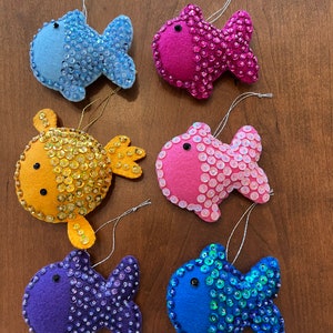 May include: Six colorful felt fish ornaments with sequin details. The ornaments are in shades of blue, pink, purple, and yellow. Each fish has a small black eye and a string for hanging. The sequins add a shimmering effect to the fish.