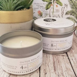 May include: Two silver candle tins with white labels. The larger tin is 8 ounces and the smaller tin is 4 ounces. Both candles are labeled "Outer Realms" and "Bigfoot Banana Bread".