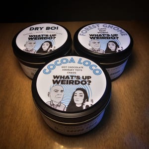 May include: Three black metal candle tins with white labels. Each label features a cartoon couple with the text "What's Up Weirdo?" and a unique scent description. The scents are "Dry Boi", "Forest Gnome", and "Cocoa Loco".