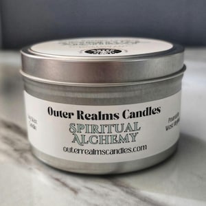 May include: A silver metal candle tin with a white label. The label reads "Outer Realms Candles" and "Spiritual Alchemy". The website address outerrealmscandles.com is also visible. The candle is on a white and gray surface.