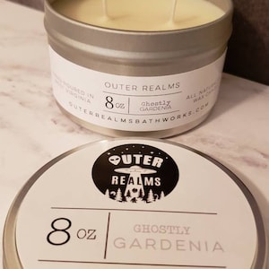 May include: An 8 ounce soy candle in a silver tin with a black and white label. The label features a UFO and the text "Outer Realms" and "Ghostly Gardenia". The candle is 100% soy and has an approximate burn time of 40 hours.