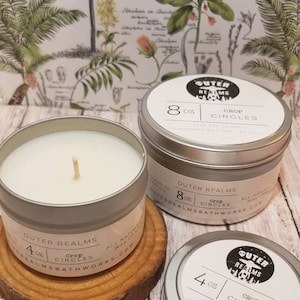 May include: Three silver-colored metal candle tins with white labels and the text "Outer Realms Crop Circles". One 4 oz candle sits on a wooden coaster, and two 8 oz candles are next to it. The background features botanical illustrations.