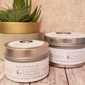 May include: Two silver-lidded candle tins with white labels, one labeled 4 oz and the other 8 oz, sit on a wooden surface. The labels read "Outer Realms" and "Sasquatch's Sandalwood." A small potted plant is in the background.