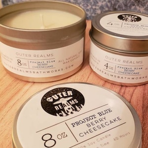 May include: Two silver candle tins with white labels. The larger tin is 8 ounces and the smaller tin is 4 ounces. Both tins are labeled "Outer Realms" and feature the text "Project Blue Berry Cheesecake". The larger tin has a black and white illustration of an alien spaceship and trees. The smaller tin has a black and white illustration of a spaceship.