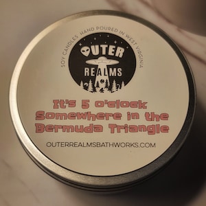 May include: A round, silver candle tin with a white label featuring the text "Outer Realms" and a graphic of a UFO over a forest. The label also reads "It's 5 o'clock Somewhere in the Bermuda Triangle."
