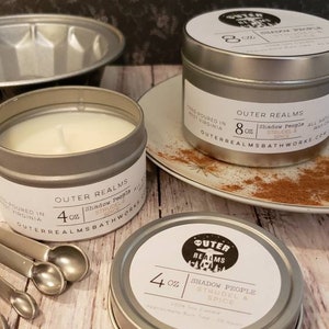 May include: Two silver candle tins with labels that read "Outer Realms" and "Shadow People Strudel & Spice". The smaller tin is 4 oz and the larger tin is 8 oz.