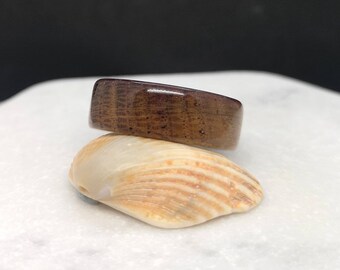 May include: A dark brown wooden ring with a natural wood grain pattern. The ring is sitting on a seashell.