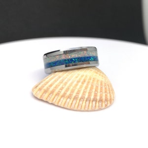 May include: A silver ring with a white, blue, and green opal inlay. The ring is sitting on a seashell.