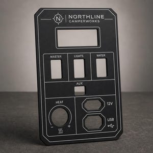 Op de afbeelding: Een zwart rechthoekig schakelpaneel met wit gegraveerde tekst en iconen. Het paneel bevat het Northline Camperworks-logo, een grote rechthoekige uitsparing, drie verticale schakelaarsleuven, een AUX-sleuf, een ronde warmteknop en 12V- en USB-poorten.