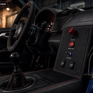 May include: A car interior featuring a black suede steering wheel with a red top marker, a manual gear shifter, and a centre console panel with a red engine start button, a master switch, toggle switches, and a blue nitrous oxide bottle.