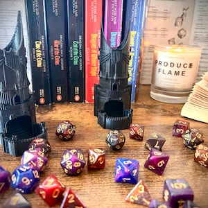 Pode incluir: Uma cena de mesa com uma torre de dados preta, vários dados poliédricos, livros e uma vela. Os dados são vermelhos, roxos, azuis e dourados. A vela diz "PRODUCE FLAME". Os livros são da série Dragonlance Legends.