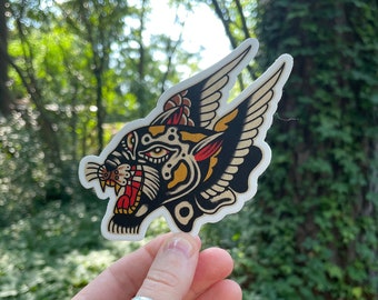 May include: A black and yellow winged panther sticker with red and white accents. The sticker is held in a hand against a green leafy background.