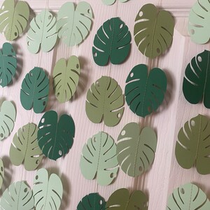 K&ouml;nnte beinhalten: Eine Girlande aus Papier-Monstera-Bl&auml;ttern in verschiedenen Gr&uuml;nt&ouml;nen, auf d&uuml;nnen Schn&uuml;ren aufgereiht. Die Bl&auml;tter haben ein detailliertes, ausgeschnittenes Design und sind vor einem hellen Hintergrund angeordnet, wodurch ein tropisches, dekoratives Thema entsteht.