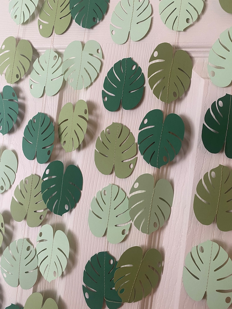 K&ouml;nnte beinhalten: Eine Girlande aus Papier-Monstera-Bl&auml;ttern in verschiedenen Gr&uuml;nt&ouml;nen, auf d&uuml;nnen Schn&uuml;ren aufgereiht. Die Bl&auml;tter haben ein detailliertes, ausgeschnittenes Design und sind vor einem hellen Hintergrund angeordnet, wodurch ein tropisches, dekoratives Thema entsteht.